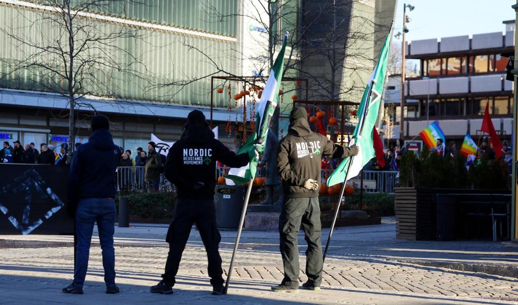 Tre menn med hettegensere som har påskriften "Nordic Resistance Movement" står bredbente på et torg med ryggen til. De holder oppe to flagg med organisasjonens logo. I bakgrunnen står en stor menneskemengde med plakater og prideflagg. Foto.