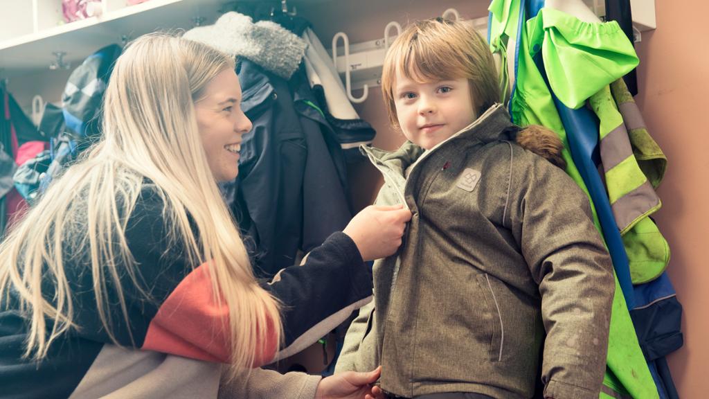 En smilende kvinnelig ansatt hjelper en gutt med å lukke glidelåsen i jakka hans i garderoben. Foto.