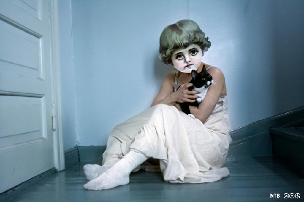 En jente med hvit kjole og dukkemaske sitter alene på gulvet og holder en svart og hvit katt. Foto.