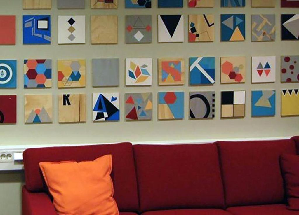 Fotografi av mange kvadratiske komposisjonar som er måla på kryssfiner og heng på ein vegg over ein sofa i eit pauserom.
