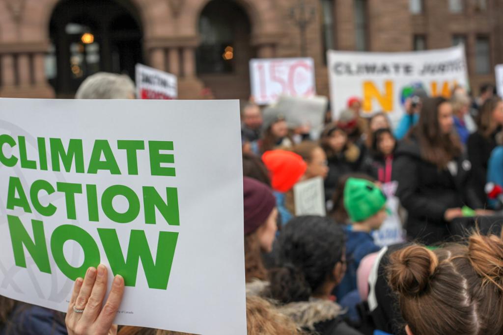En plakat hvor det står skrevet "climate action now" i forgrunnen, og i bakgrunnen ser man mange mennesker på en klimamarkering. Foto.