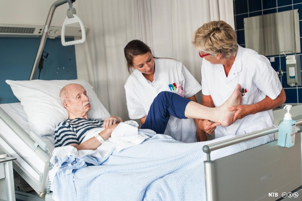 To helsefagarbeidere undersøker foten til en eldre, sengeliggende pasient. Foto.