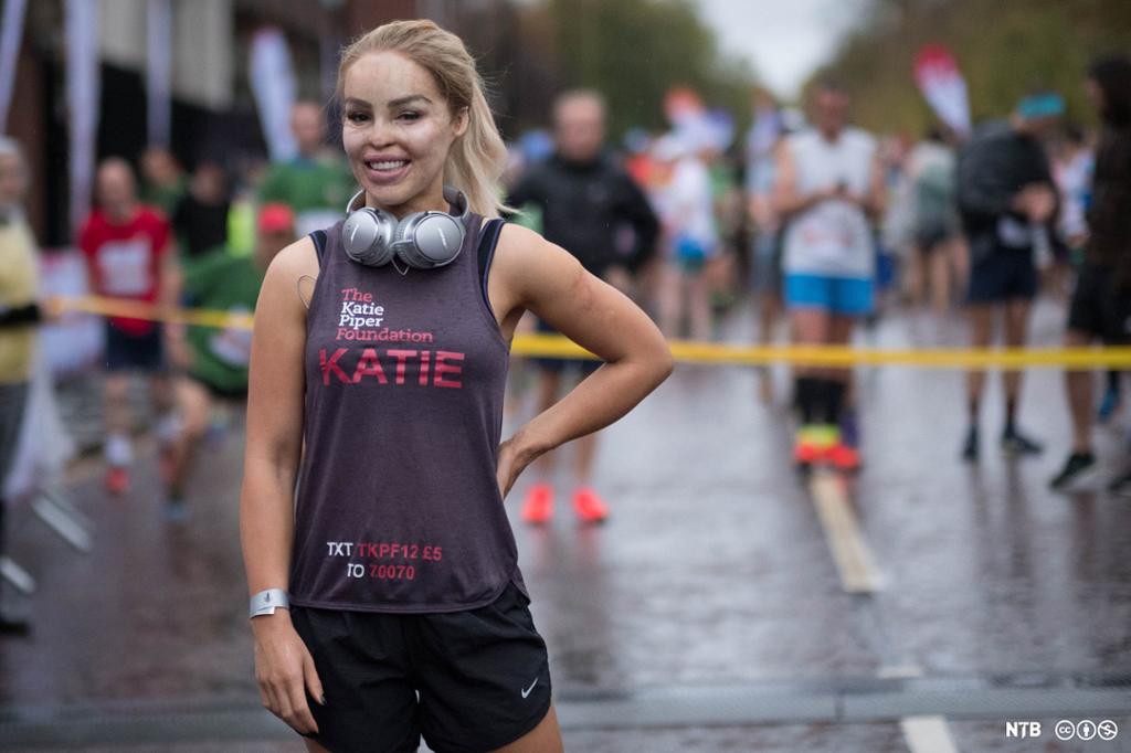 Woman, Katie Piper, in training gear. Behind her is a yellow ribbon accross the road, the background is blurred but we can see runners standing there. 