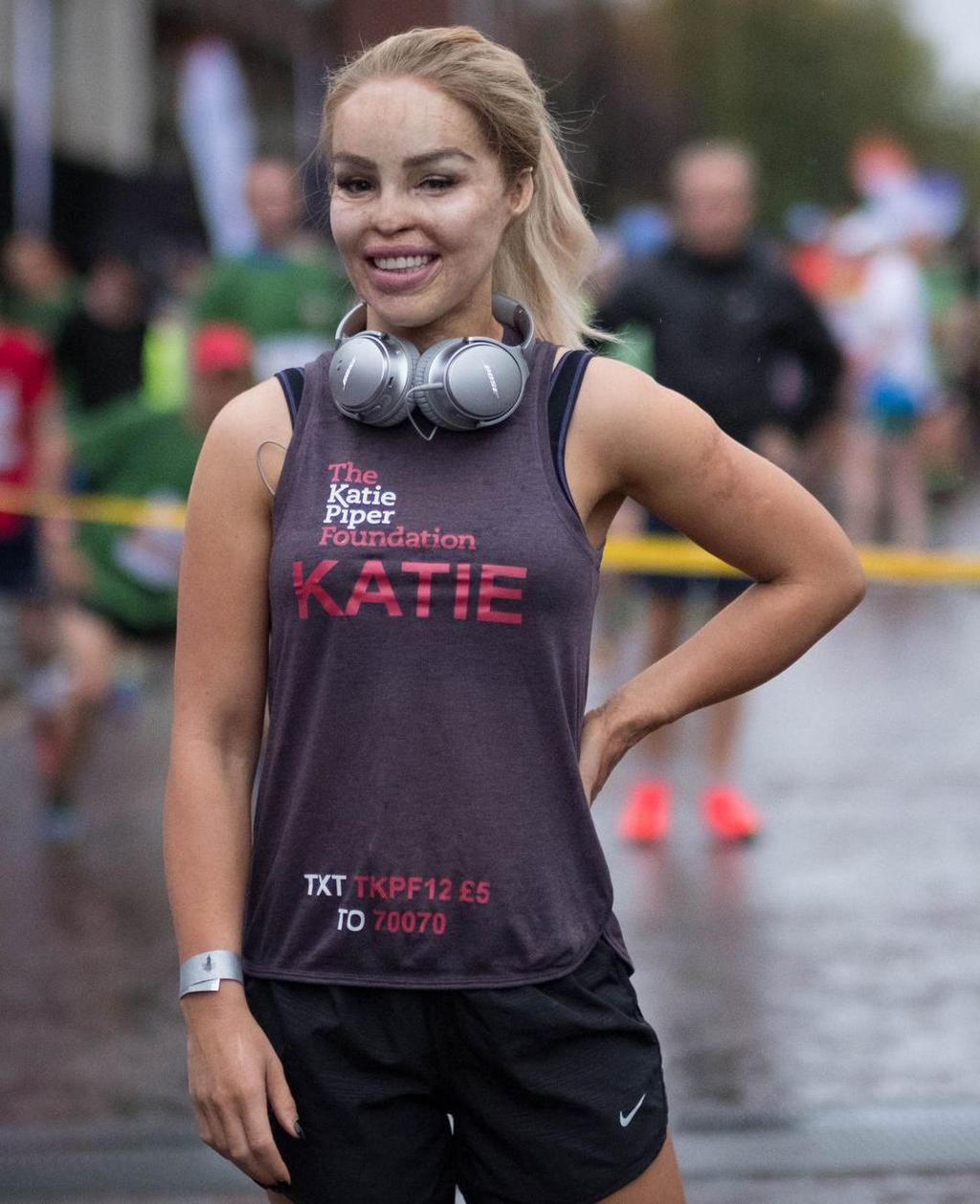 Woman, Katie Piper, in training gear. Behind her is a yellow ribbon accross the road, the background is blurred but we can see runners standing there. Photo.