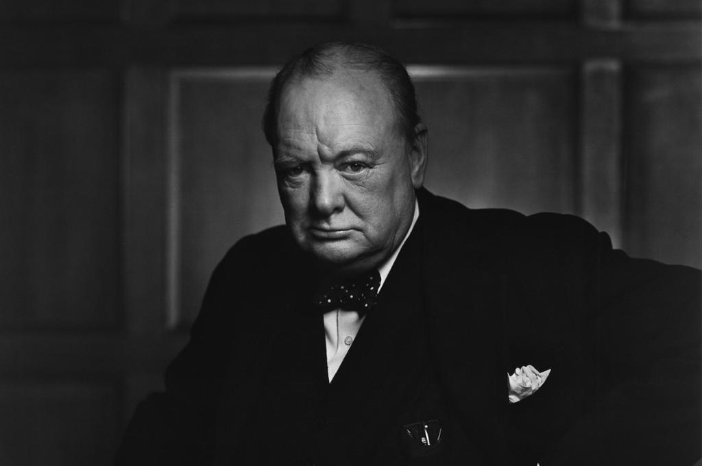 Portrait of Winston Churchill holding a cane. We see a stout, older man. He is wearing a dark jacket, bow tie, and a white shirt: He has a pocket handkerchief. He looks serious. Photo.