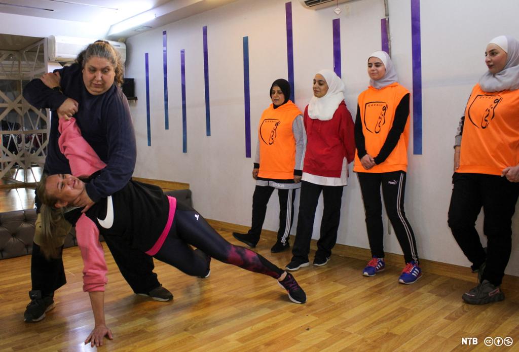 En instruktør vise hvordan man foretar et halsgrep mens fire unge kvinner i treningsbukser og hijab følger med. Foto.