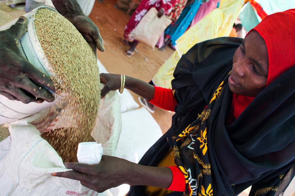 Black woman in red head scarf holds bag open as a bucket of grain is poured into it. 