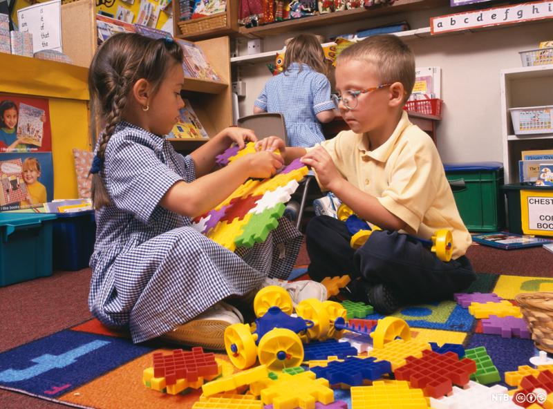 Ei jente og en gutt sitter sammen på gulvet og bygger med store, fargerike plastbiter som kan settes sammen. I bakgrunnen sitter ei jente ved en pult. Foto.