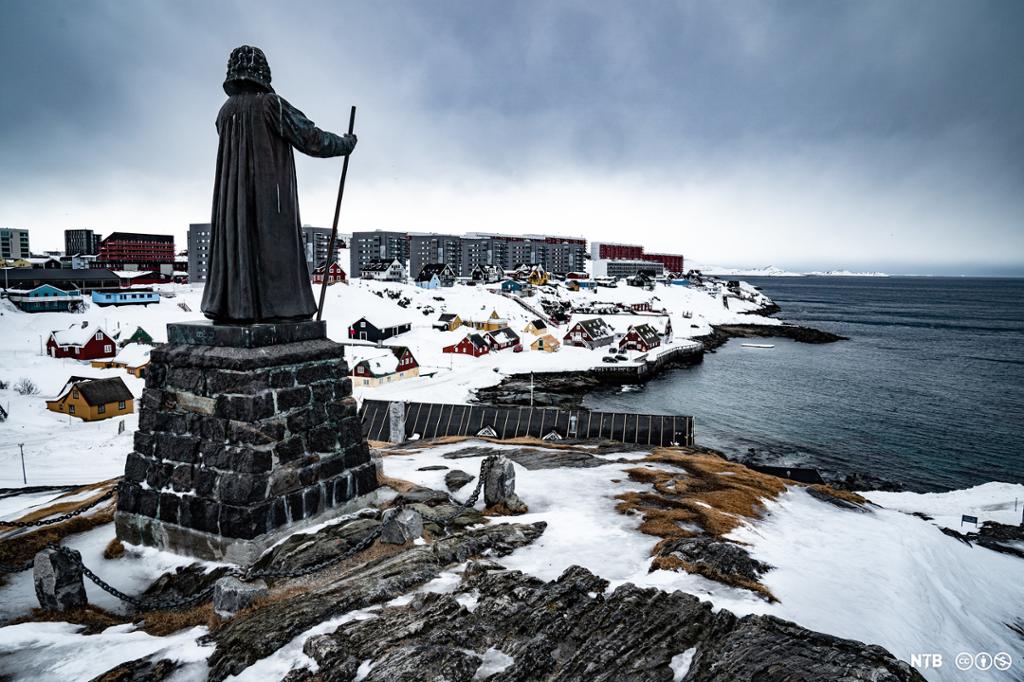 Statue av Hans Egede som held ein stav i Nuuk, hovudstaden på Grønland. Han står på ei lita høgde med utsikt mot havet. Tradisjonelle hus og moderne bygningar i bakgrunnen. Foto. 