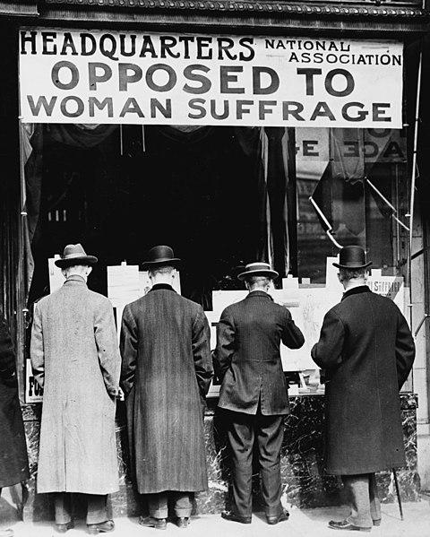 A black and white photo showing four men from behind standing outside a building marked with the sign "Headquarters Opposed to Woman Suffrage." 