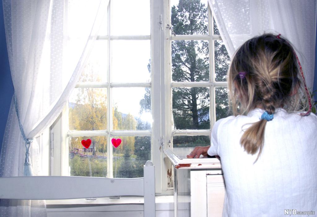 Ung jente sitter ved vinduet på rommet sitt. Det står to små røde hjerter i vinduskarmen. Foto.