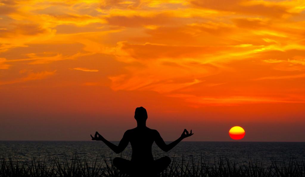Person mediterer i solnedgang. Personen sitter på ei strand med ryggen til og armene halvveis opp. Hen ser på sjøen og sola som stiger opp fra havet. Foto.