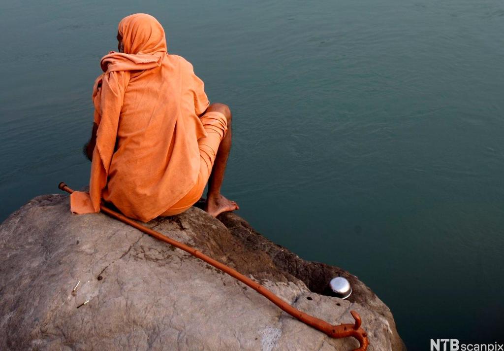 Mann med oransje kappe sit ved ei elv. Foto.