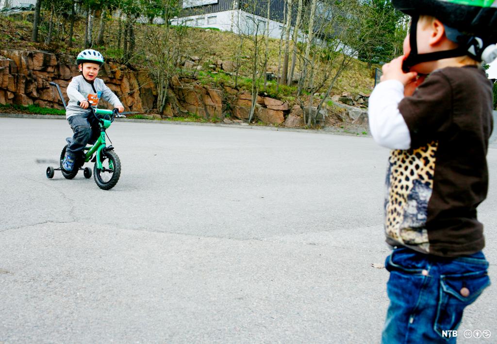 En gutt sitter på en grønn sykkel med støttehjul. Han har på seg sykkelhjelm. En annen gutt holder på å feste sykkelhjelmen, han har ingen sykkel. Foto.