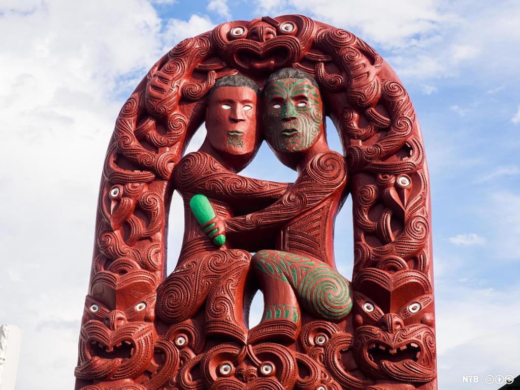 Photo: We see a large carved wood sculpture of two seated people. They have their arms against each other's torsos and their knees are touching. Their eyes are painted white. They are surrounded by grotesque faces with white staring eyes. The sculpture is mostly red, but there are also parts that are green. 