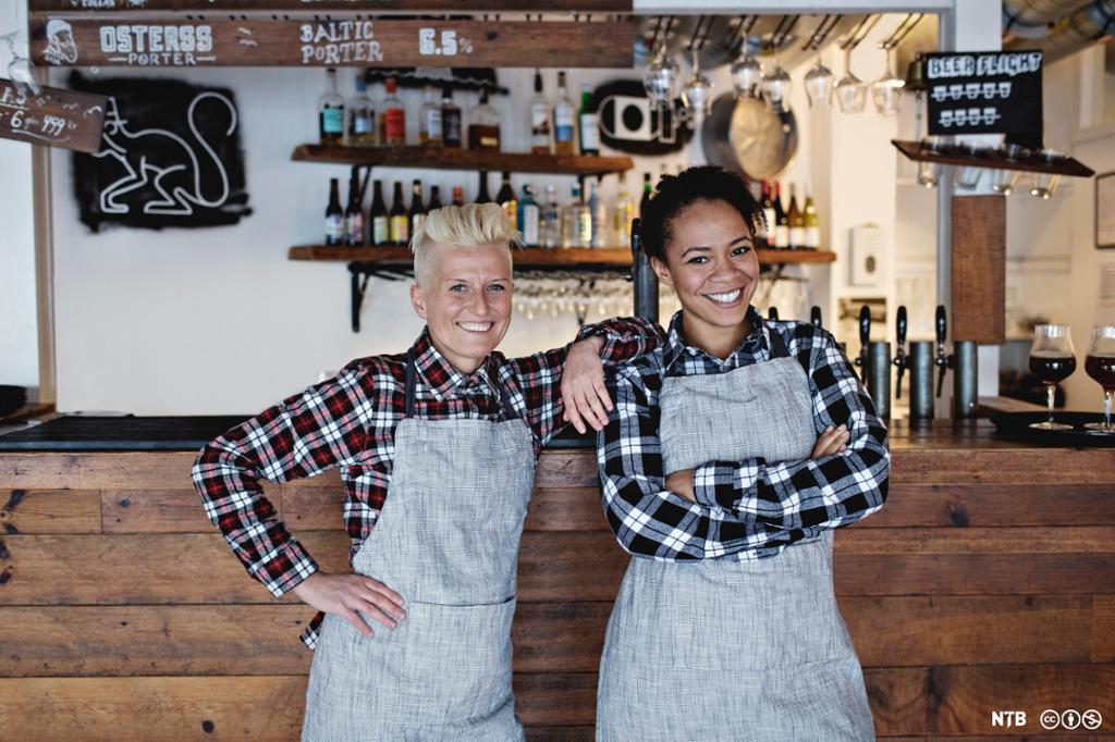 To smilende kvinnelige servitører står lent mot en bardisk. Foto.