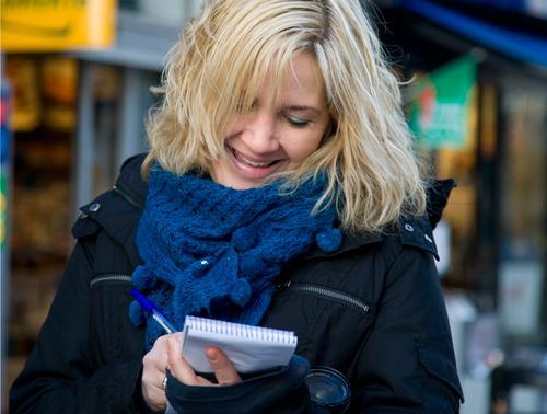En kvinne med jakke og skjerf står i en gate og noterer på en notisblokk. Foto.