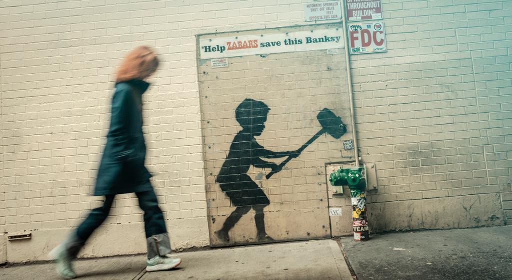 The street art graffiti, called ‘Hammer Boy’, is a piece by Banksy. It depicts a child with a hammer, poised to smash a fire hydrant. Above the artwork, the text reads, ‘Help ZABAR save this Banksy.’ A blurred person can be seen walking past the mural. Photo.