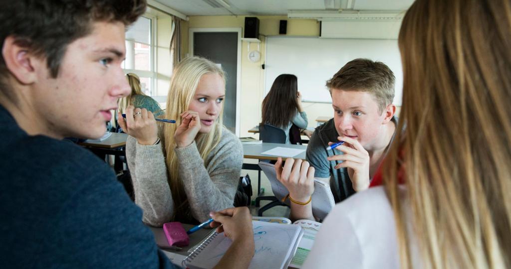 Foto av fire ungdommer som diskuterer rundt en pult i et klasserom.