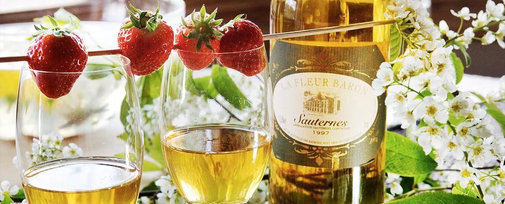 En flaske med Sauternes-vin, to vinglass fylt med vinen, og noen friske jordbær stukket på pinne ligger over glassene. Foto.