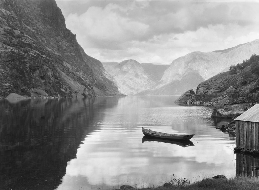 Robåt fortøydd ved eit naust i ein fjord omkransa av høge fjell. Svart-kvitt-foto. 