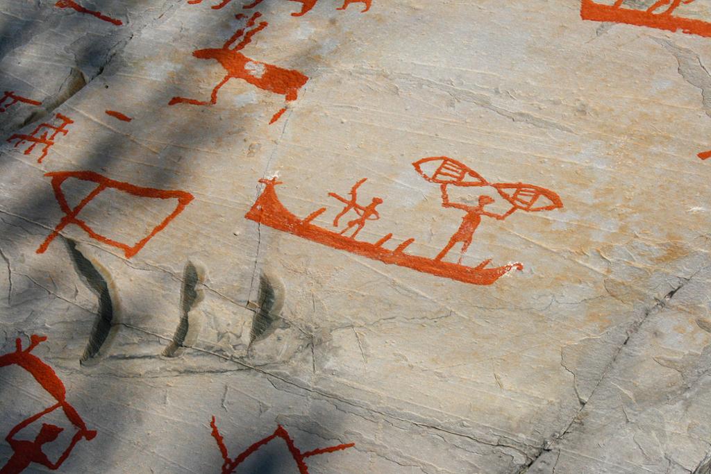 Utsnitt av helleristingar måla i raudt på grått fjell. Utsnittet viser menneske i båt, samt dyr ved sida av. Foto.