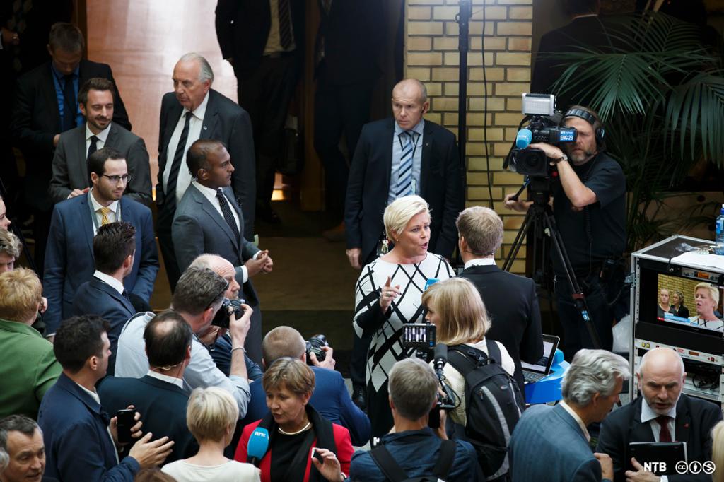 Daværende finansminister Siv Jensen står i hvit kjole omgitt av journalister. Foto.