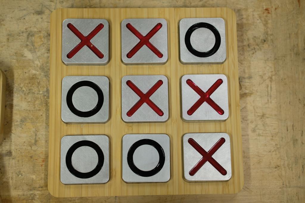 Tre-på-rad-spel med firkanta bambusbrett og firkanta brikker av aluminium. Brikkene er merkte med eit stort raudt kryss eller ein svart sirkel. Foto.