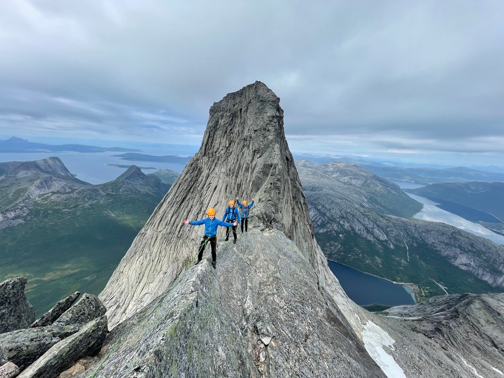 Tre personar iførte klatreutsyr står på toppen av eit veldig høgt fjell. Foto.