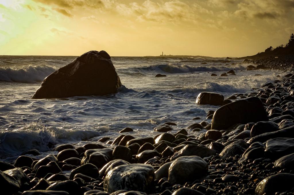 Bilde av steiner ved en strand og bølger som er på vei inn på stranda. Til venstre i bildet vises en stor stein i vannet. I bakgrunnen skimtes et fyrtårn. Foto.