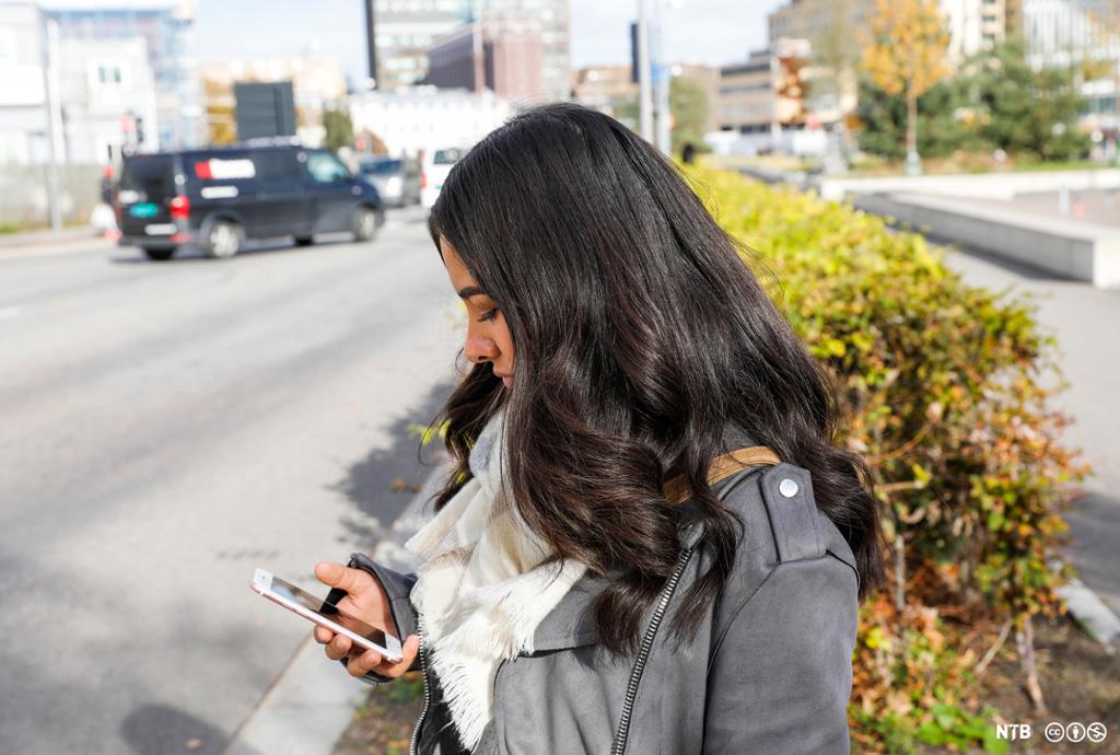 Nærbilde av jente på 17–18 år med langt, mørkt hår. Ho står og ser på mobilen sin. Jenta har på seg ei grå skinnjakke og eit kvitt skjerf. 