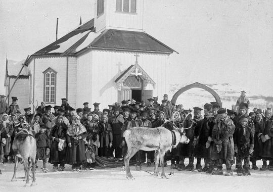 En stor gruppe personer, en del i samiske drakter, og noen reinsdyr, foran en kirke. Bildet er tatt vinterstid. Svart-hvitt foto. 