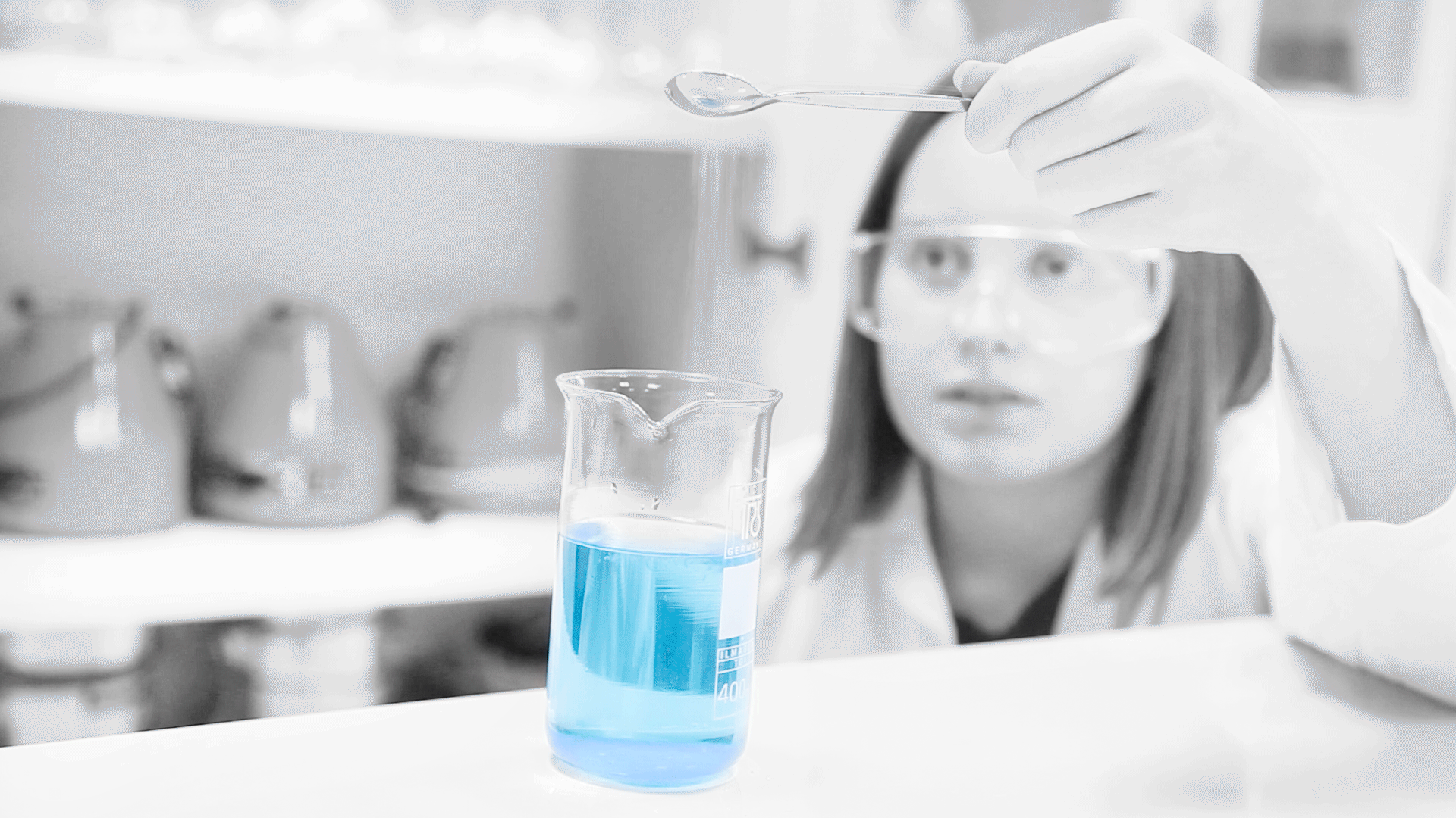 En jente heller salt fra en skje over i en blå væske i et begerglass. Foto.