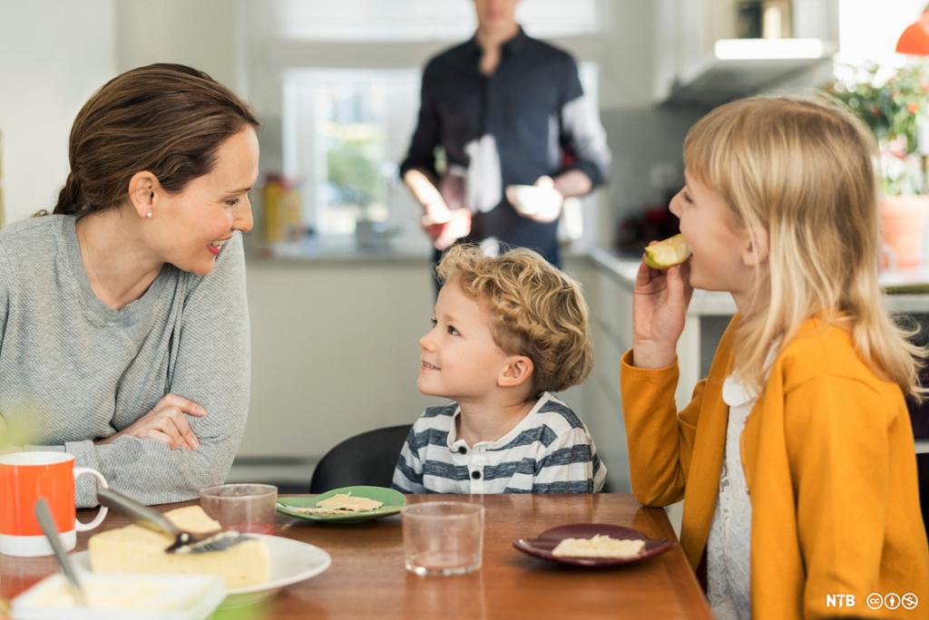 En voksen kvinne, en liten gutt og ei litt større jente spiser frokost. I bakgrunnen står en mann som holder to små skåler i handa. Foto.