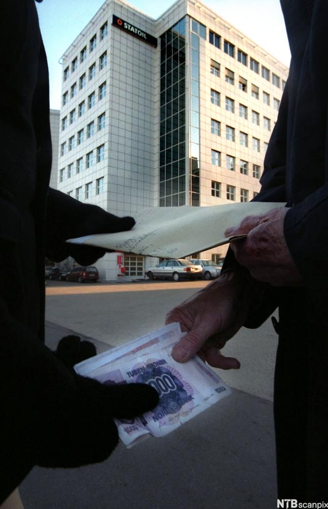 Ein mørkkledd person rekkjer ein konvolutt til ein annan mørkkledd person, som rekkjer ein bunke tusenlappar tilbake. I bakgrunnen ser vi eit kontorbygg med Statoil-logo. Foto.