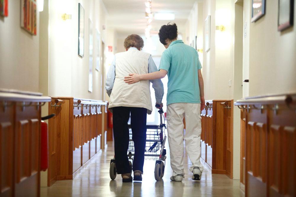 En helsefagarbeider holder en støttende hånd på ryggen til en eldre kvinne med rullator mens de går bortover en gang. Foto.