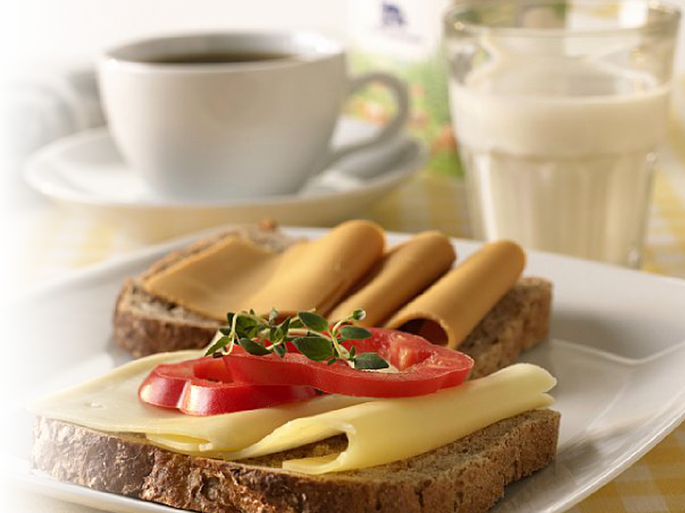 To grove brødskiver på en tallerken, en med gulost og paprika, en med brunost. Et glass melk og en kaffekopp i bakgrunnen. Foto.