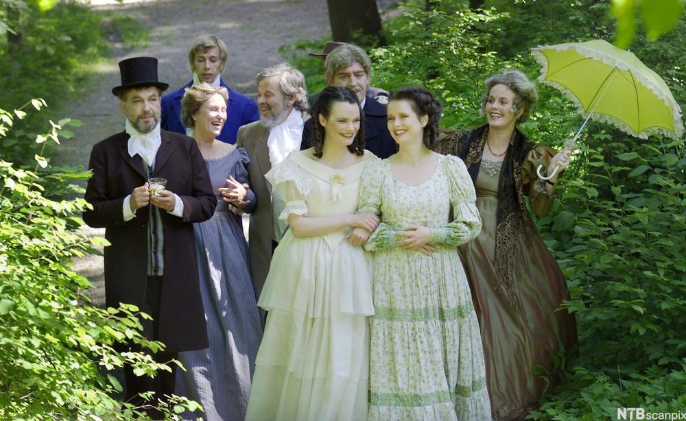 Dramatisering av Camilla Colletts "Amtmannens døtre ". Flere unge mennesker pynta i fine klær spaserer i en hage. Foto.