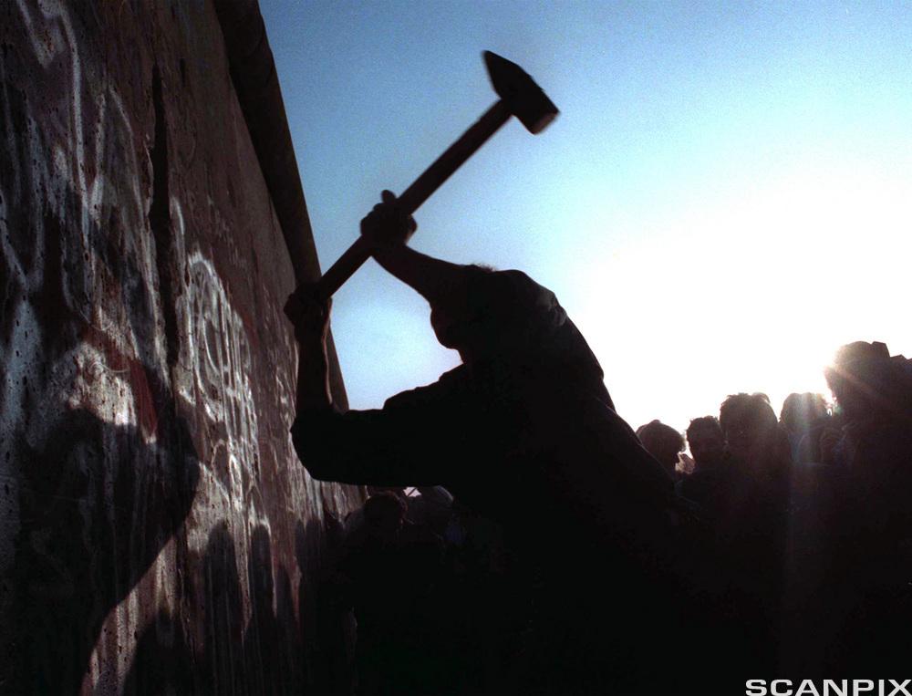 En person svinger en diger hakke mot en mur. I bakgrunnen står en folkemengde. Foto.
