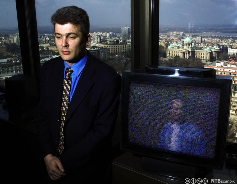 Mann i dress står foran et TV-apparat med forstyrrelser på skjermen på grunn av dårlig tv-signal. Foto.