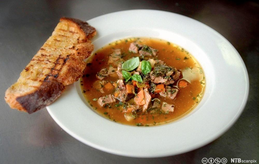 En tallerken med suppe med kjøttbiter. Basilikumblader på toppen og et stykke ristet brød ved siden av. Foto.