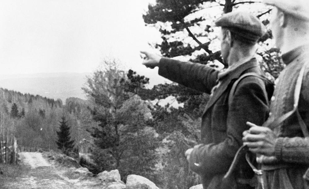 To menn med ryggsekk langs en skogsvei. Den ene mannen peker mot åsene i bakgrunnen. Svart-hvitt foto.