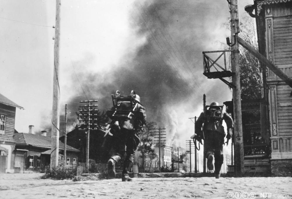 Soldater løper gjennom ei gate  mellom brennende hus. Foto.
