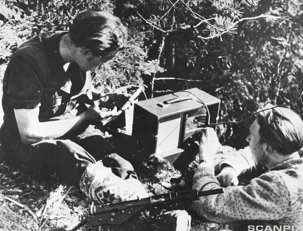 To menn lytter til et radioapparat ute i skogen. Foto.