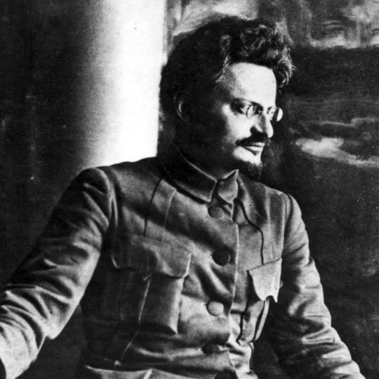 Lev Trotskij iført uniformsjakke med brystlommer. Han har kort, mørkt hår og briller. Foto.