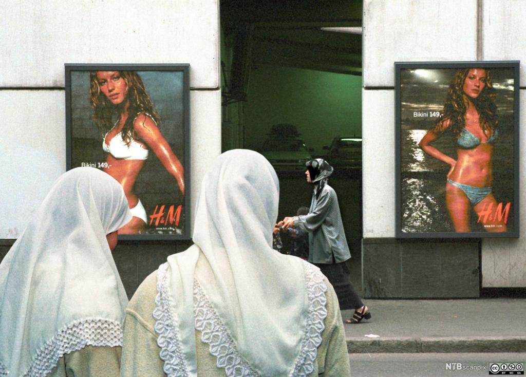 Muslimske kvinner med slør foran reklameplakater med lettkledde vestlige modeller. Foto.