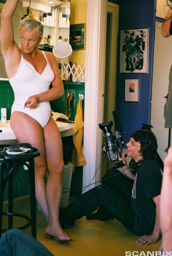 En mann med grått hår står iført kvinneundertøy på et baderom. På gulvet sitter en yngre mann med videokamera. Foto.