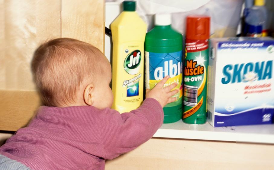 Baby griper etter en flaske vaskemiddel som står i et kjøkkenskap. Foto.