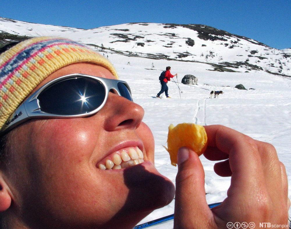 Kvinne spiser appelsin i strålende sol på påskefjellet. Hun har på solbriller og lue og er brun i huden. I bakgrunnen ses en kvinne med hund som går på ski. Foto.