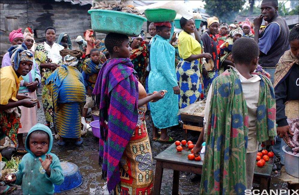 Det deles ut mat i en menneskemengde. Kvinner med fargerike afrikanske klær bærer ting på hodet. Et barn i møkkete klær spiser. Foto.